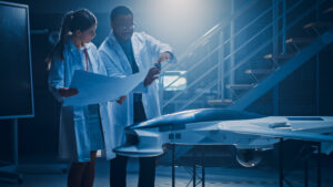 Two Aerospace Engineers Work On Unmanned Aerial Vehicle / Drone Prototype. Aviation Scientists in White Coats Holding Blueprints. Laboratory with Commercial Aerial Surveillance Aircraft